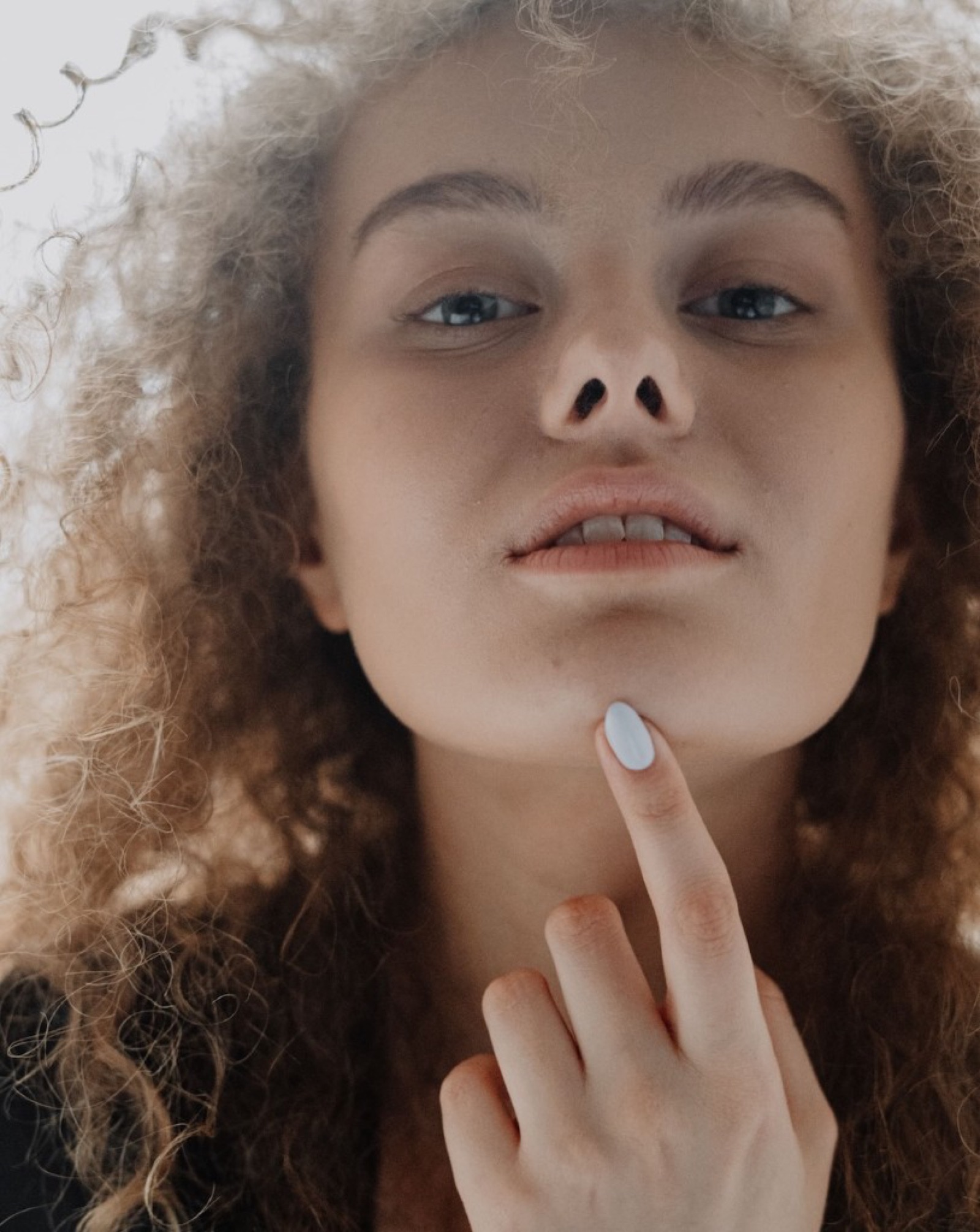 Close-up of a woman with curly blonde hair resting a finger on her chin against a bright background